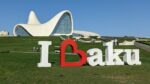A sign that says I "heart" Baku. The heart shape is turned sideways and forms the B of Baku. It sits on a large green lawn, and behind it is the Heydar Aliyev Centre, a large curvy white edifice with large windows.
