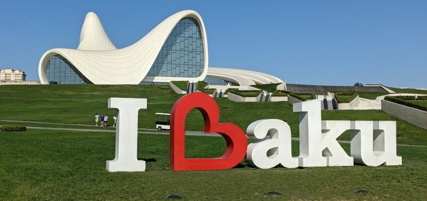 A sign that says I "heart" Baku. The heart shape is turned sideways and forms the B of Baku. It sits on a large green lawn, and behind it is the Heydar Aliyev Centre, a large curvy white edifice with large windows.