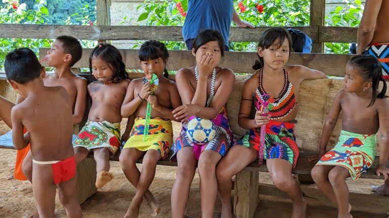 A group of indigenous children sitting on a bench wearing traditional costumes